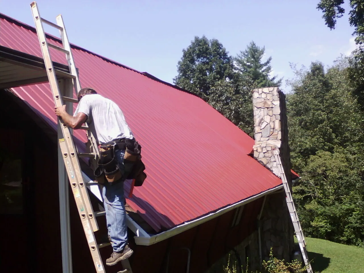 Roofer on a ladder beside a red metal roof and stone chimney — Soffit & Fascia Repair by Roofing Brothers in Mansfield Center
